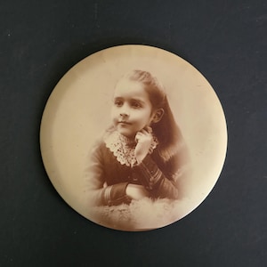 May include: A sepia-toned portrait of a young girl, set within a circular frame. She is wearing a dark dress with a lace collar and her long hair is styled to the side. The image has a vintage aesthetic.