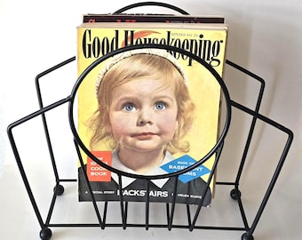 May include: A black wire magazine rack holding vintage magazines. The magazines have a yellow cover with the words "Good Housekeeping" and a picture of a young girl. The rack has a handle and rests on four small feet.