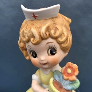 May include: A vintage ceramic figurine of a nurse with blonde curly hair, wearing a white cap with a red cross and a yellow dress. The figurine is holding a small pot with a flower and blue leaves.