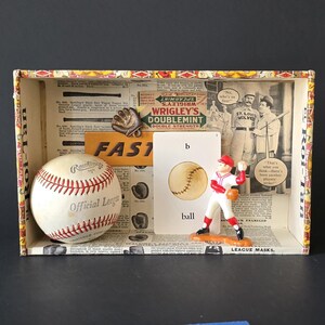 May include: A baseball-themed display box with a vintage aesthetic. It contains a baseball, a baseball glove, a baseball player figurine, and a card with the word "ball". The box also features newspaper clippings and the word "FAST".
