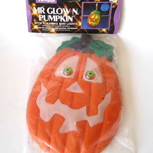 May include: Orange Mr. Glow N. Pumpkin Halloween decoration with flashing mini lights. The pumpkin has a carved face with white accents and green eyes. The product is packaged in a clear plastic bag with a purple and black header.