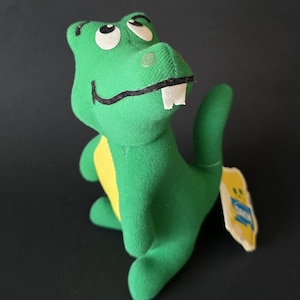 May include: A green plush alligator toy with a yellow belly and white teeth. The toy has white eyes and a black smile. A yellow tag with blue text is attached to the toy.