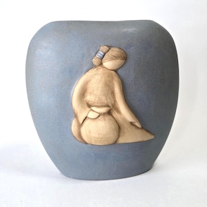 May include: A blue ceramic vase with a matte finish. A beige relief sculpture of a person in a robe is on the front. The person is seated and holding a round object. The vase has a rounded shape.