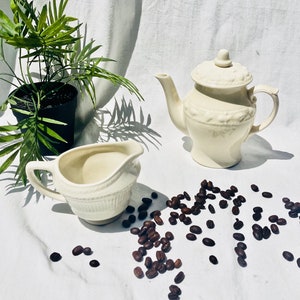 May include: A white ceramic teapot and creamer set with a floral design. The teapot has a lid and a spout. The creamer has a handle. Coffee beans are scattered around the set.