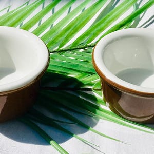 May include: Two small, brown ceramic ramekins with white interiors and a brown rim. The ramekins are set on a white surface with green palm leaves. Ideal for serving individual portions of food.
