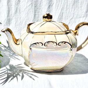 May include: Cream-colored teapot with gold accents. The teapot features a pearlescent finish, a gold-trimmed lid, handle, and spout. Decorative scrollwork adorns the front. A small gold crown sits atop the lid. The teapot is a vintage item.