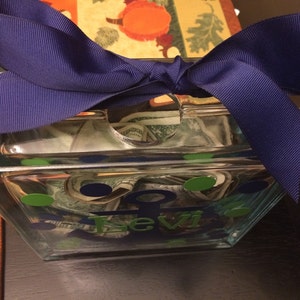 May include: A clear glass bank with a navy blue ribbon tied on top. The bank is decorated with green and blue oval stickers and the name "Levi" in blue lettering. The bank is filled with rolled-up paper money.