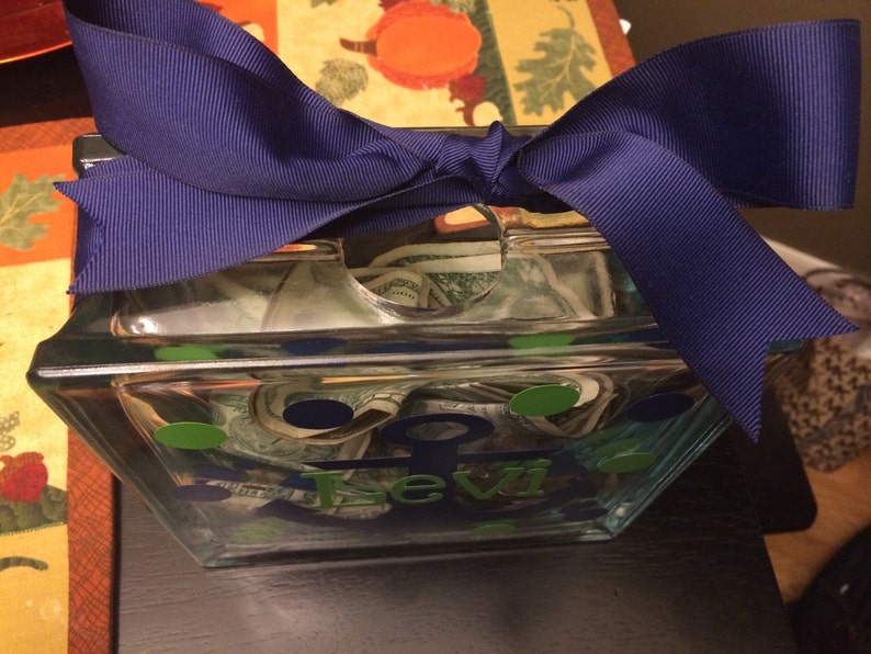 May include: A clear glass bank with a navy blue ribbon tied on top. The bank is decorated with green and blue oval stickers and the name "Levi" in blue lettering. The bank is filled with rolled-up paper money.