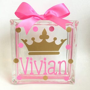 May include: A clear glass block decorated with a gold crown, pink polka dots, and the name "Vivian" in pink. A large pink bow sits atop the block, creating a decorative gift item.