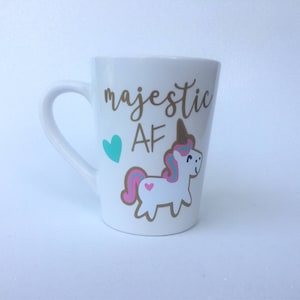 May include: White ceramic mug with a gold unicorn and the text "majestic AF" in gold lettering. A teal heart is to the left of the text.