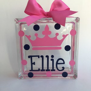 May include: A clear glass block decorated with a pink crown, navy blue and pink polka dots, and the name "Ellie" in navy blue. A large pink bow sits atop the block, adding a decorative touch.