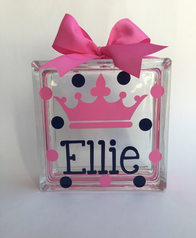 May include: A clear glass block decorated with a pink crown, navy blue and pink polka dots, and the name "Ellie" in navy blue. A large pink bow sits atop the block, adding a decorative touch.