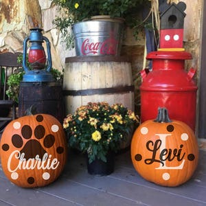 May include: Two decorated pumpkins with the names "Charlie" and "Levi" on them. The pumpkins are sitting on a wooden porch with a red milk can, a wooden barrel, and a lantern in the background.
