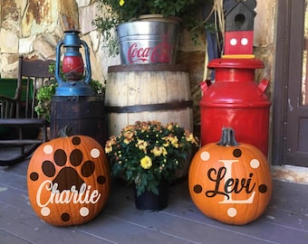 May include: Two decorated pumpkins with the names "Charlie" and "Levi" on them. The pumpkins are sitting on a wooden porch with a red milk can, a wooden barrel, and a lantern in the background.