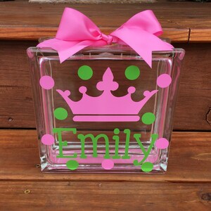 May include: A clear glass block decorated with a pink crown, green and pink polka dots, and the name "Emily" in green. A large pink bow sits atop the block. This decorative block is a personalized gift.