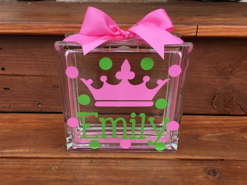 May include: A clear glass block decorated with a pink crown, green and pink polka dots, and the name "Emily" in green. A large pink bow sits atop the block. This decorative block is a personalized gift.