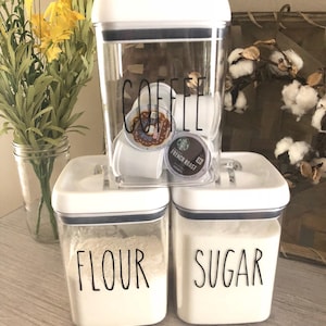 May include: Three white square canisters with black lids and labels. The top canister is labeled "COFFEE" and contains coffee pods. The middle canister is labeled "FLOUR" and the bottom canister is labeled "SUGAR".