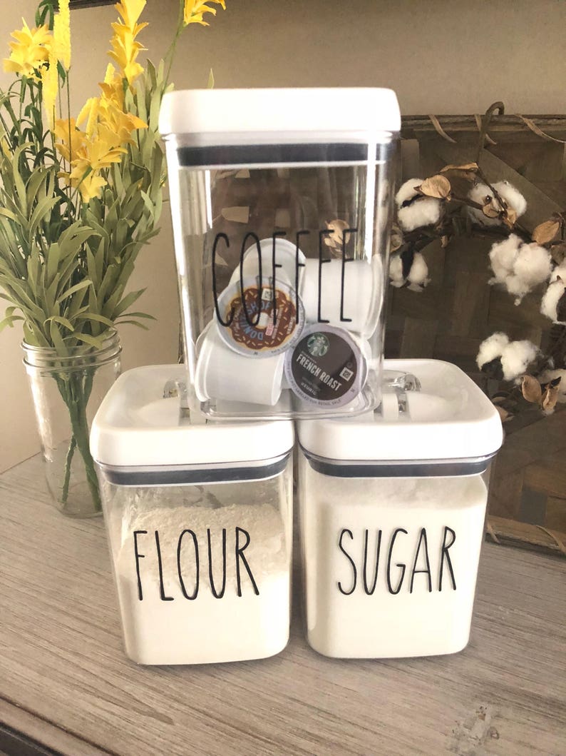 May include: Three white square canisters with black lids and labels. The top canister is labeled "COFFEE" and contains coffee pods. The middle canister is labeled "FLOUR" and the bottom canister is labeled "SUGAR".