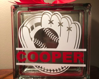 Puede incluir: Un bloque de vidrio con un diseño de béisbol y guante blanco y el nombre "COOPER" en letras rojas. El bloque está decorado con un lazo rojo.