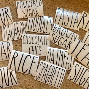 May include: A collection of white labels with black handwritten text for various pantry items, including "Flour", "Sugar", "Rice", "Pasta", "Tea", "Coffee", "Oatmeal", "Spaghetti", "Macaroni Noodles", "Chocolate Chips", "Brown Sugar", and "Powdered Sugar".