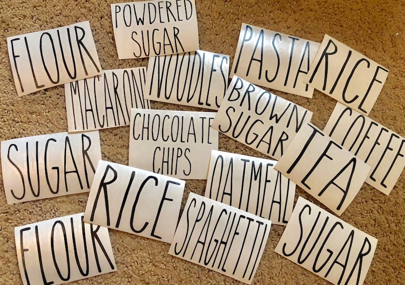 May include: A collection of white labels with black handwritten text for various pantry items, including "Flour", "Sugar", "Rice", "Pasta", "Tea", "Coffee", "Oatmeal", "Spaghetti", "Macaroni Noodles", "Chocolate Chips", "Brown Sugar", and "Powdered Sugar".