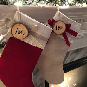 May include: Two red and white Christmas stockings with wooden ornaments that say "Ava" and "Levi" hanging on a white mantel.