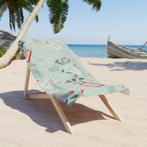 May include: A light blue beach chair with a botanical pattern featuring red accents and text. The chair is made of wood and fabric, set on a sandy beach with a boat in the background.