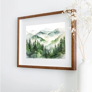 Puede incluir: Pintura a la acuarela de un paisaje monta&ntilde;oso con &aacute;rboles de hoja perenne y valles brumosos. La obra de arte est&aacute; enmarcada en un marco de madera marr&oacute;n oscuro y se exhibe en una pared blanca. La pintura presenta tonos de verde y azul.