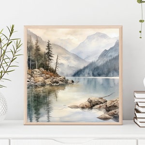 Op de afbeelding: Een aquarel schilderij van een bergmeer sc&egrave;ne met een mistige achtergrond. Het meer wordt omringd door bomen en bergen, en er zijn rotsen op de voorgrond. Het schilderij is ingelijst in een licht houten lijst.