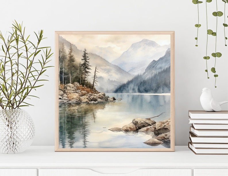 Op de afbeelding: Een aquarel schilderij van een bergmeer sc&egrave;ne met een mistige achtergrond. Het meer wordt omringd door bomen en bergen, en er zijn rotsen op de voorgrond. Het schilderij is ingelijst in een licht houten lijst.