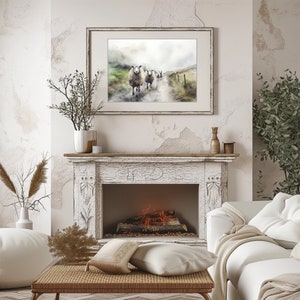 May include: A watercolor painting of a group of sheep walking down a path in a misty landscape. The painting is framed in a white frame and hangs above a white fireplace with a lit fire.