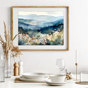 May include: A watercolor painting of a misty mountain landscape with a foreground of blue and green trees and wildflowers. The mountains are painted in shades of blue and gray, with a soft, hazy effect. The painting is framed in a light wood frame.