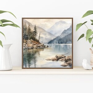 Op de afbeelding: Een aquarel schilderij van een bergmeer sc&egrave;ne met een klein eiland op de voorgrond. Het meer is omringd door bergen en bomen, en het water is lichtblauw. Het schilderij is ingelijst in een bruin frame.