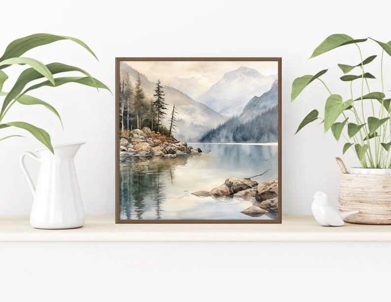 Op de afbeelding: Een aquarel schilderij van een bergmeer sc&egrave;ne met een klein eiland op de voorgrond. Het meer is omringd door bergen en bomen, en het water is lichtblauw. Het schilderij is ingelijst in een bruin frame.