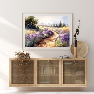 May include: A watercolour painting of a lavender field with a dirt path leading through it. The painting is framed and hanging on a white wall above a wooden cabinet with a wire mesh front.