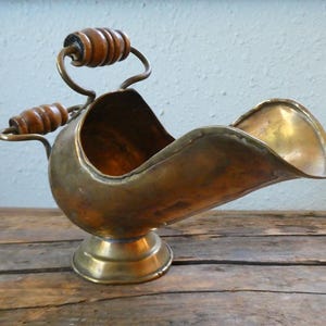 May include: A brass coal scuttle with a polished metallic finish, featuring a wide, open-mouthed scoop design, a circular pedestal base, and two curved metal handles adorned with dark, ribbed wooden grips for handling the vessel.