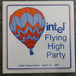 May include: A square white tile with a colorful hot air balloon design. The balloon is orange and blue, with the Intel logo and the words "Flying High Party." The text "Intel Corporation April 27, 1987" is also present.