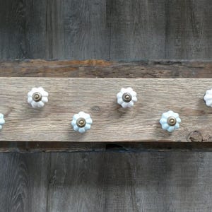 May include: A rustic wooden coat rack with six white and light blue floral knobs. The rack is made of reclaimed wood and has a natural, weathered appearance. The knobs have a brass-colored center.