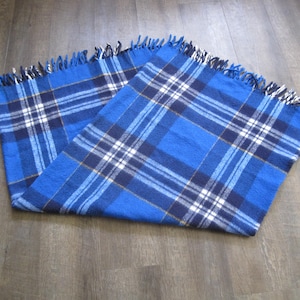 May include: A blue plaid throw blanket with fringe on all sides. The blanket is folded in half and laid flat on a wooden floor.