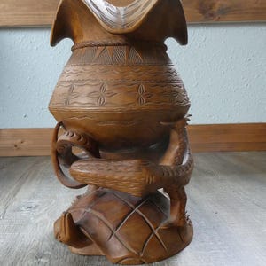 May include: A brown carved wooden vessel featuring a wide, flared rim and a rounded body decorated with geometric patterns. The base is supported by a sculpted turtle figure with a textured shell, while a dragon-like creature wraps around the center.