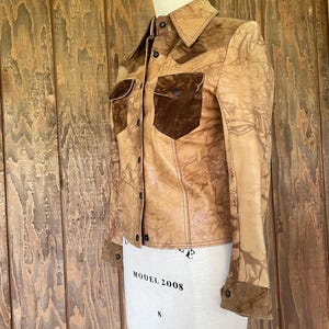 Peut inclure: Une veste en cuir marron avec un col et des poches en daim. La veste a un aspect vieilli avec un motif marbr&eacute;.