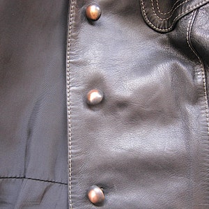 May include: Close-up of a black leather jacket with silver buttons and white stitching. The jacket has a smooth, textured surface.