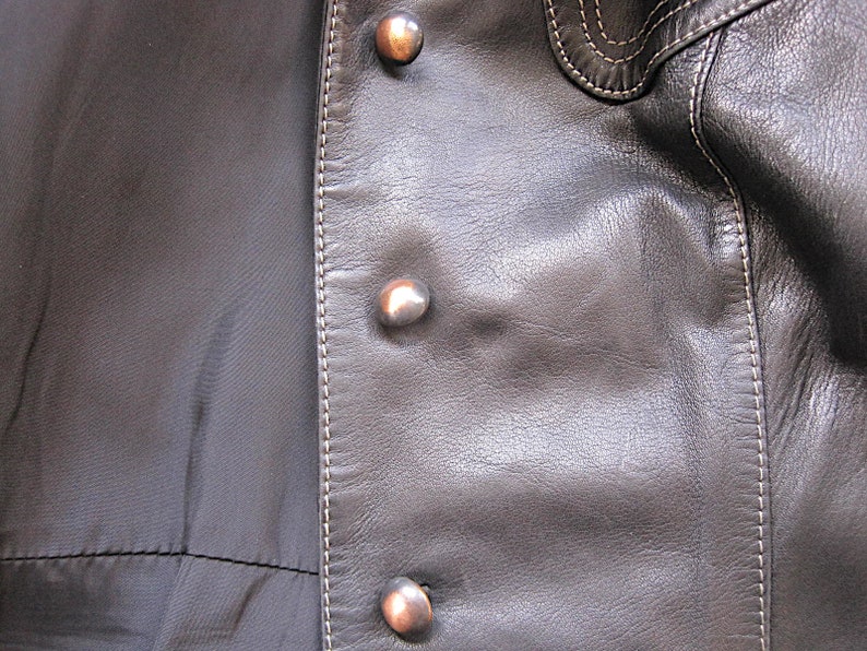 May include: Close-up of a black leather jacket with silver buttons and white stitching. The jacket has a smooth, textured surface.