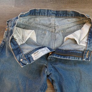 May include: Blue denim Wrangler jeans with the zipper partially open. The jeans have a classic five-pocket design, with a visible label. The interior pockets are white, and the stitching is visible.