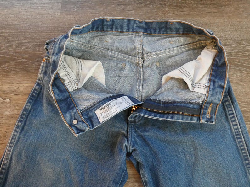 May include: Blue denim Wrangler jeans with the zipper partially open. The jeans have a classic five-pocket design, with a visible label. The interior pockets are white, and the stitching is visible.