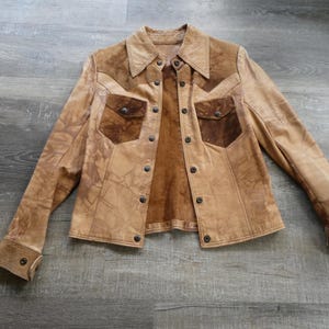 Peut inclure: Une veste en cuir marron avec des accents en daim et un devant &agrave; boutons. La veste a un aspect vieilli et comporte deux poches &agrave; rabat sur la poitrine.