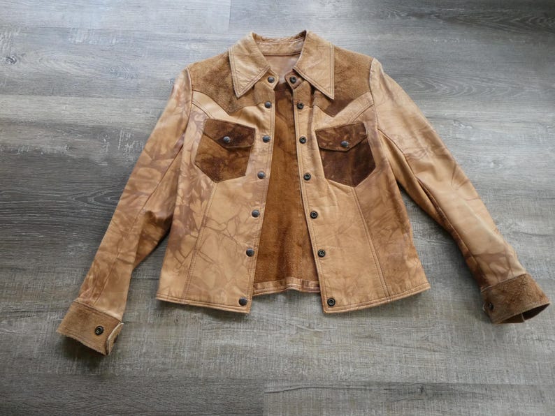 Peut inclure: Une veste en cuir marron avec des accents en daim et un devant &agrave; boutons. La veste a un aspect vieilli et comporte deux poches &agrave; rabat sur la poitrine.