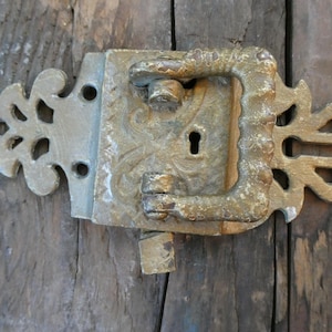 May include: Antique metal door latch with a keyhole and ornate handle. The latch is a weathered gold color with decorative floral accents. The latch is mounted on a weathered wooden door.
