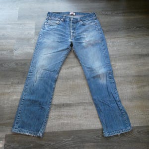May include: A pair of light blue denim jeans featuring a classic five-pocket design, a button-fly closure, and a faded wash with whiskering details at the hips. The jeans have a straight-leg cut with visible stitching and a red brand tag on the pocket.