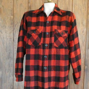 May include: A red and black plaid flannel shirt with two chest pockets and button-up closure. The long-sleeved shirt is displayed on a mannequin against a wooden background. The shirt has a classic lumberjack style.